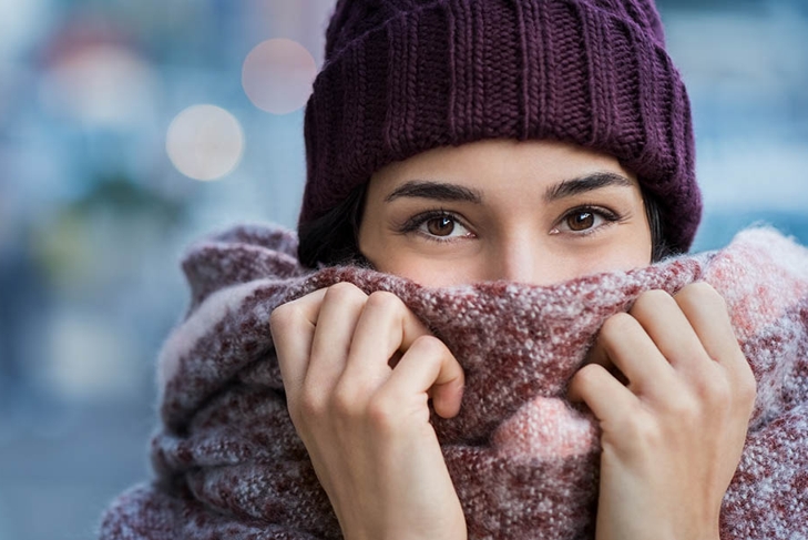 Winter portrait of young beautiful woman covering face with woolen scarf. Closeup of happy girl feeling cold outdoor in the city. Young woman holding scarf and looking at camera.